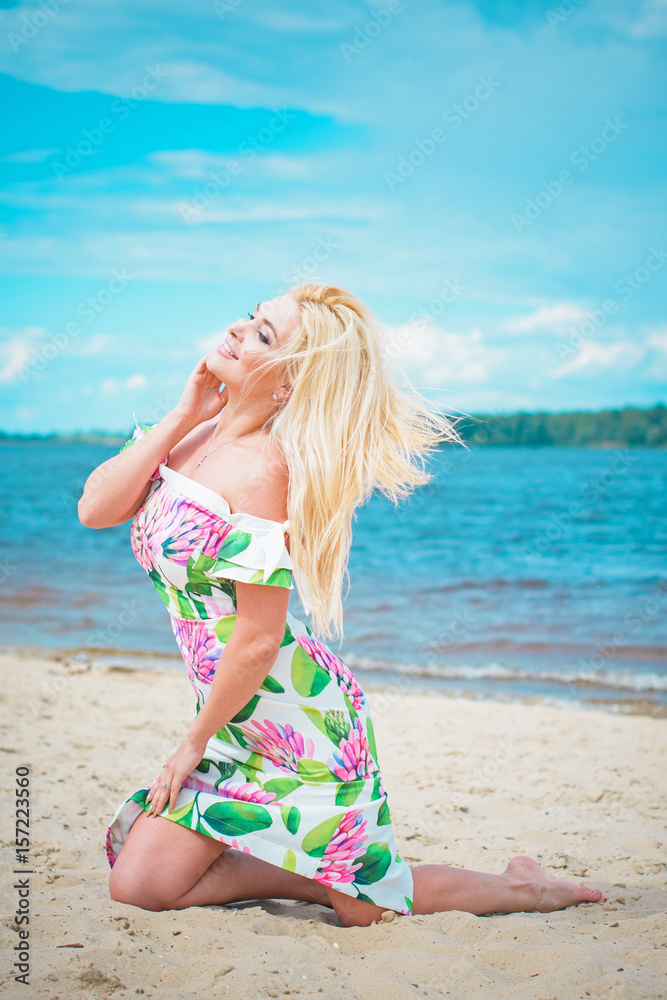 Beautiful blonde hair woman in romantic flowers dress near blue water and sand. Cute happy girl have fun on a nature