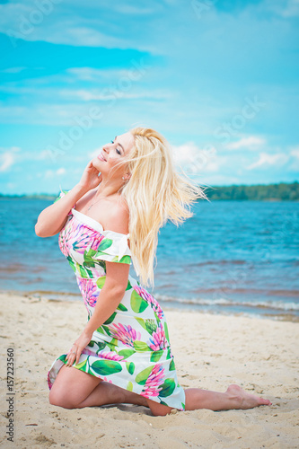 Beautiful blonde hair woman in romantic flowers dress near blue water and sand. Cute happy girl have fun on a nature