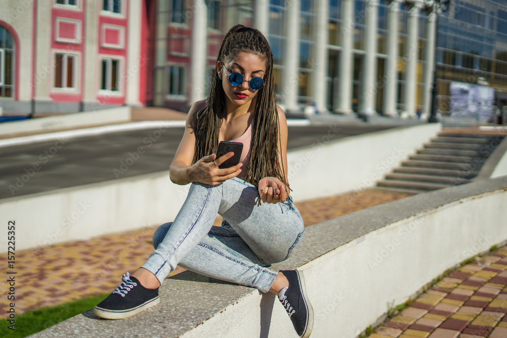 Fototapeta premium beautiful girl with zizi cornrows dreadlocks chatting on her smartphone. Urban street style
