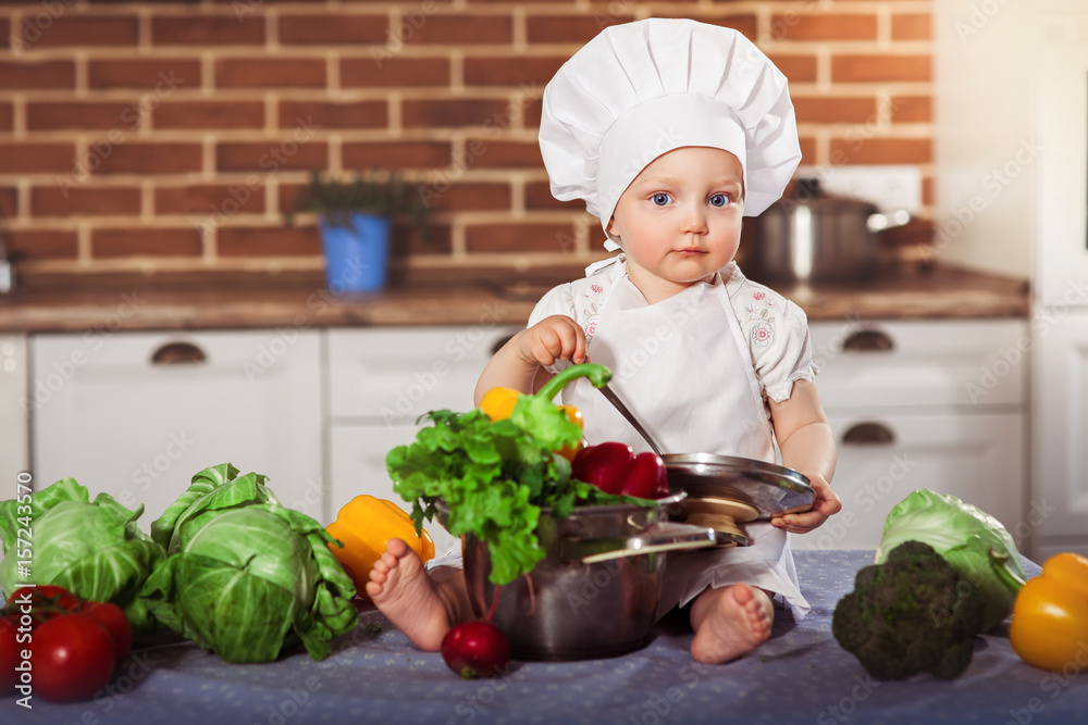 Baby girl dressed in white chef toque and apron sits with spoon near stainless pan filled by raw vegetables. Brick wall background. One year old child nutrition. Horizontal Baby girl dressed in white chef toque and apron sits with spoon near stainless pan filled by raw vegetables. Brick wall background. One year old child nutrition. Horizontal