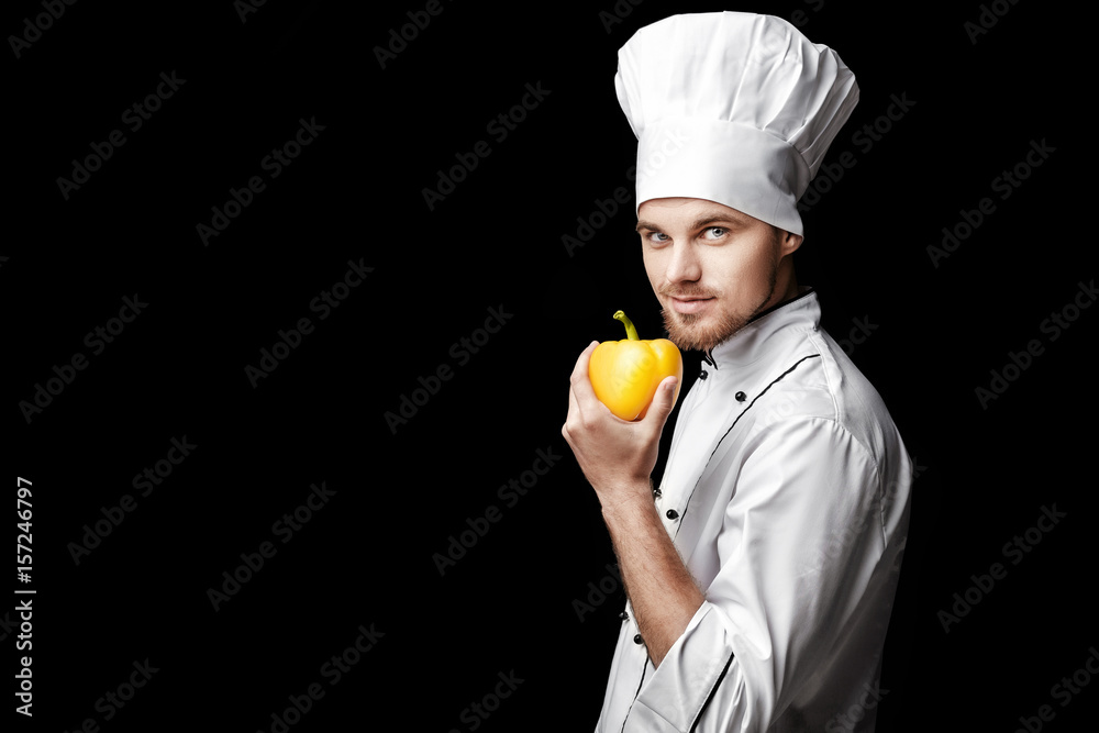 Young bearded man chef In white uniform holds yellow bell pepper on black background Young bearded man chef In white uniform holds yellow bell pepper on black background