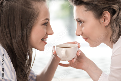 young smiling girlfriends drinking coffee and looking at each other