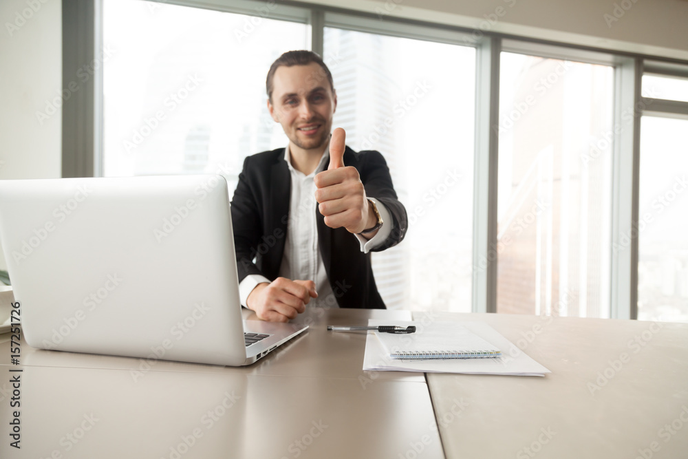 Confident businessman showing thumbs up gesture at desk in office. Entrepreneur satisfied with work results. CEO supports good decision, recommends proven solution. Company leader like successful idea