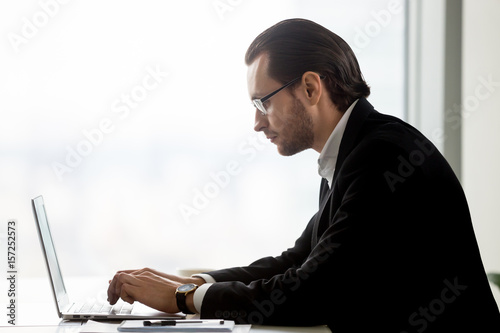 Side view portrait of stylish businessman in glasses typing on laptop. Successful entrepreneur working on company marketing strategy, analyzes new markets. CEO focused on work, writing annual plan