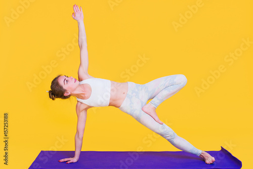 Young yogini woman stretching on purple mat isolated on yellow background