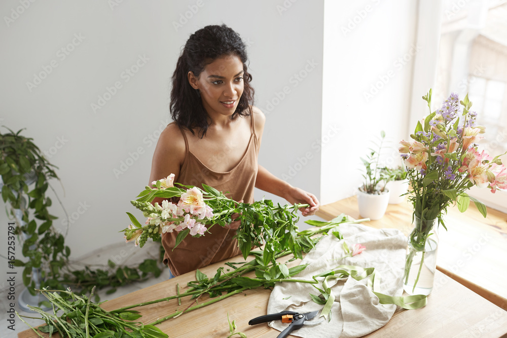 Young beautiful african girl florist taking care of flowers at workplace over white wall. Young beautiful african girl florist taking care of flowers at workplace over white wall.