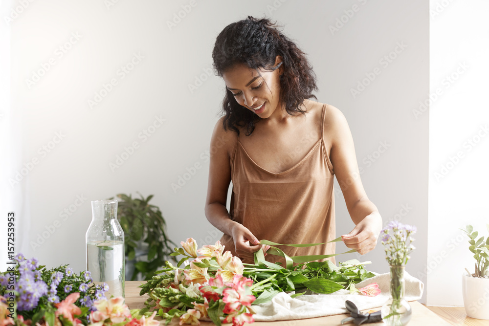 Cheeful african girl florist smiling holding ribbon working with flowers over white wall. Cheeful african girl florist smiling holding ribbon working with flowers over white wall.