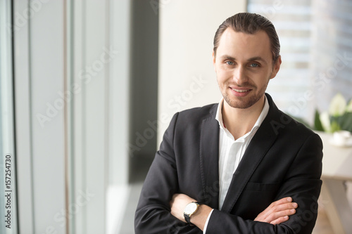 Portrait of young successful company leader standing with arms crossed and looking in camera with smile. Entrepreneur confident in his own abilities posing in office interior. Satisfied by success CEO