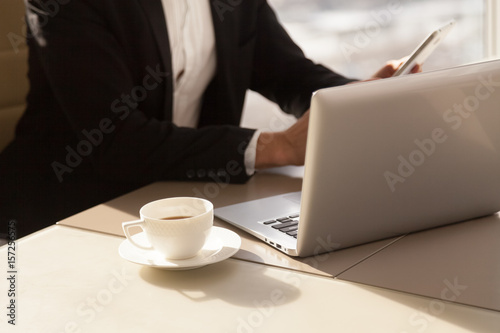 Businessman using mobile phone at desk with laptop during early coffee break in office. Entrepreneur working with electronic devices at workplace. Cup of refreshing coffee or tea at morning, close up