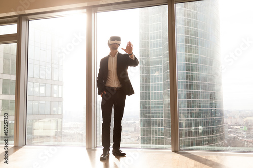 Businessman in VR headset enjoys 3d visualization of apartment or office interior design. Man in virtual reality glasses standing with raised hand near skyscraper window with urban landscape outside