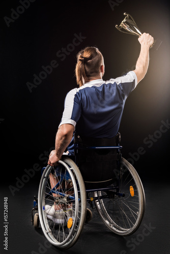 Fotografie rear view of paralympic sitting in wheelchair and holding champion goblet isolat
