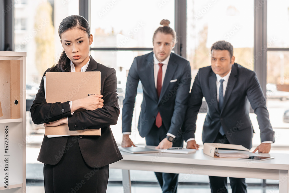 Stressed young businesswoman holding folder and businessmen standing behind Stressed young businesswoman holding folder and businessmen standing behind