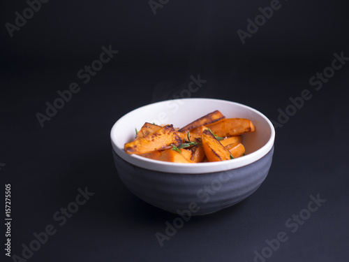 Fried sweet potatoes wedges sprinkled with wild thyme and sea salt, served in rustic grey and white bowl on black background 