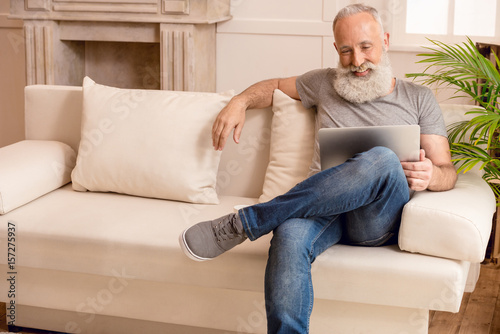senior bearded man smiling and using laptop while sitting on sofa at home © LIGHTFIELD STUDIOS