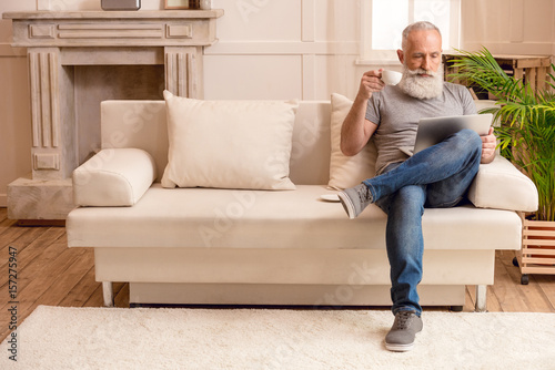 senior bearded man drinking coffee and looking on laptop while sitting on sofa at home