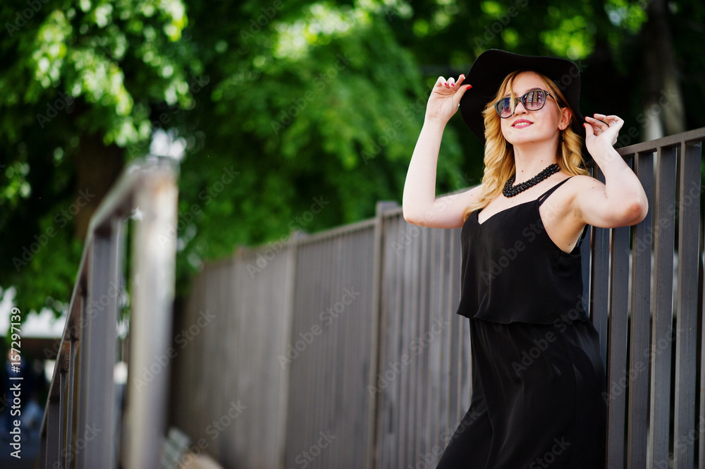 Blonde woman on black dress, sunglasses, necklaces and hat posed at streets.