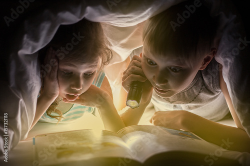 Children bedtime. Sister and brother are reading a book under a blanket with flashlight. Pretty young boy and lovely girl having fun in children room.