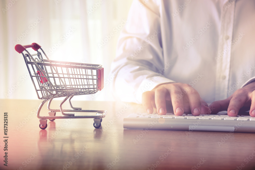 Online shopping concept. Man's hands typing on computer keyboard next to a shopping cart. Concept for background, website banner, promotional materials, presentation templates, advertising. Online shopping concept. Man's hands typing on computer keyboard next to a shopping cart. Concept for background, website banner, promotional materials, presentation templates, advertising.