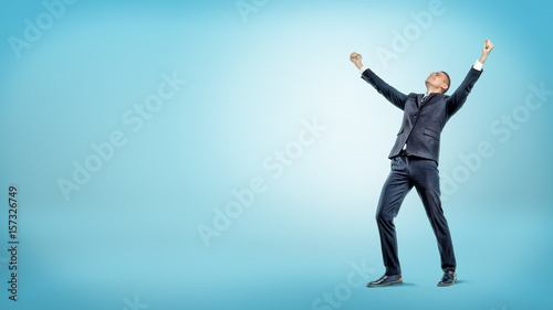 A joyful businessman standing with hands raised in victory and looking up on blue background.