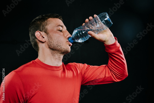 Profile portrait of attractive young European sportsman drinking water from bottle looking up wearing red sportswear, relaxing after run or workout. Sport motivation. Lifestyle.