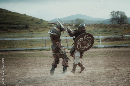 Russia, Feodosiya, May 9, 2017, Medieval joust knights in helmets and chain mail battle on swords with shields in their hands, reconstruction. European Middle Ages Performance festival