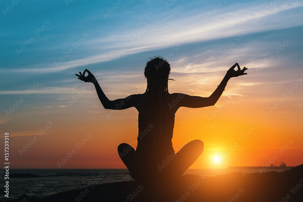 Silhouette of young yoga woman does meditation on sea beach at amazing sunset.