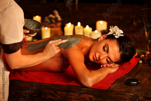 Mud mask of woman in spa salon. Back massage with clay full body . Girl on luxary interior with candles in oriental therapy room. Female lying on red towel in wooden spa bed.
