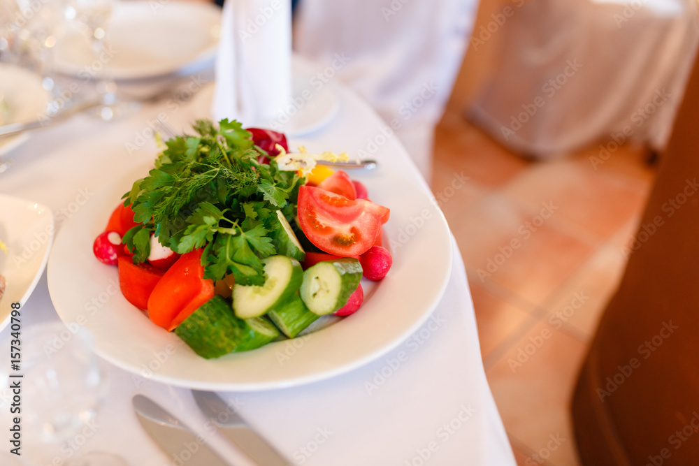 Delicious summer salad with fresh vegetables on a white plate served on the table . Cucumber, tomatoes, radishes, and lettuce.