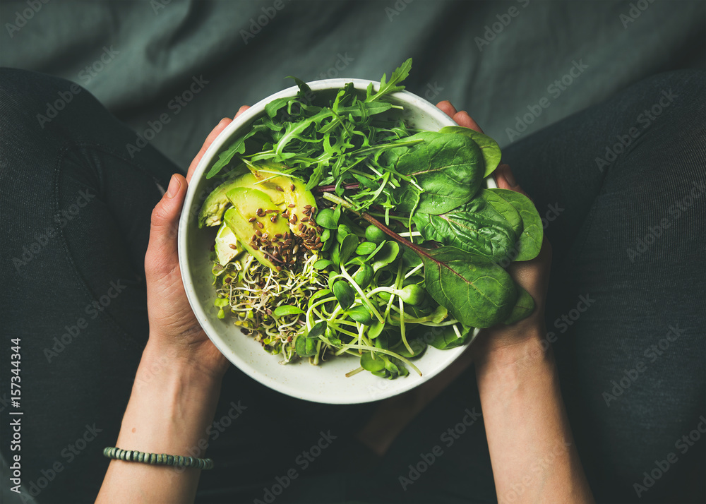 Green vegan breakfast meal in bowl with spinach, arugula, avocado, seeds and sprouts. Girl in leggins holding plate with hands visible, top view. Clean eating, dieting, vegan food concept - obrazy, fototapety, plakaty 