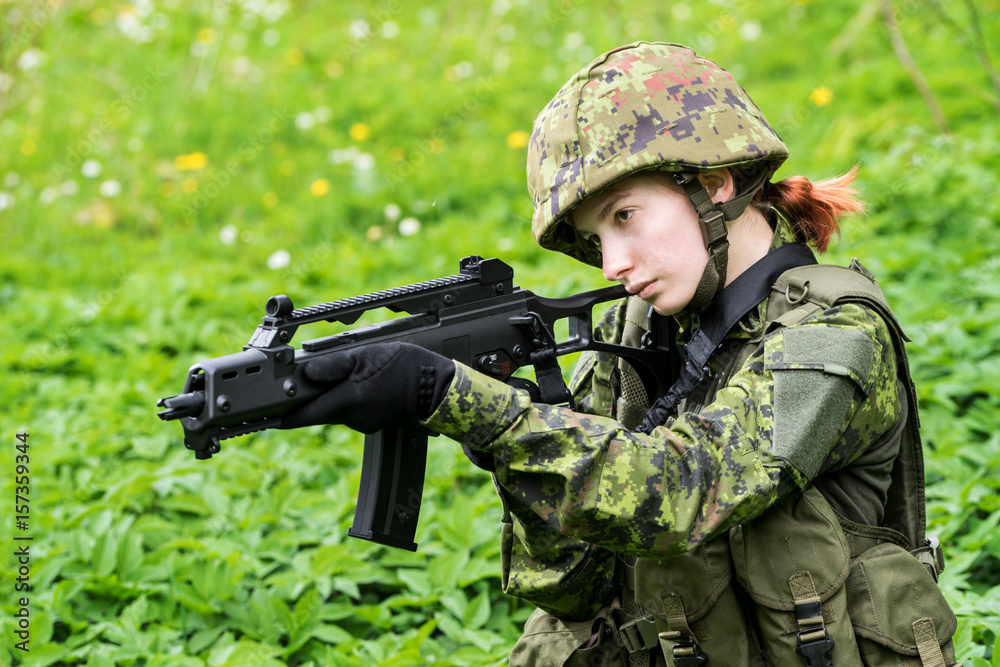 Portrait of armed woman with camouflage. Young female soldier observe with firearm. Child soldier with gun in war, green goutweed background.  Military, army people concept