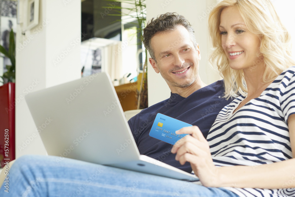 They like to shop online. Shot of a smiling middle aged couple sitting on couch and using credit card and laptop while shopping online. They like to shop online. Shot of a smiling middle aged couple sitting on couch and using credit card and laptop while shopping online.