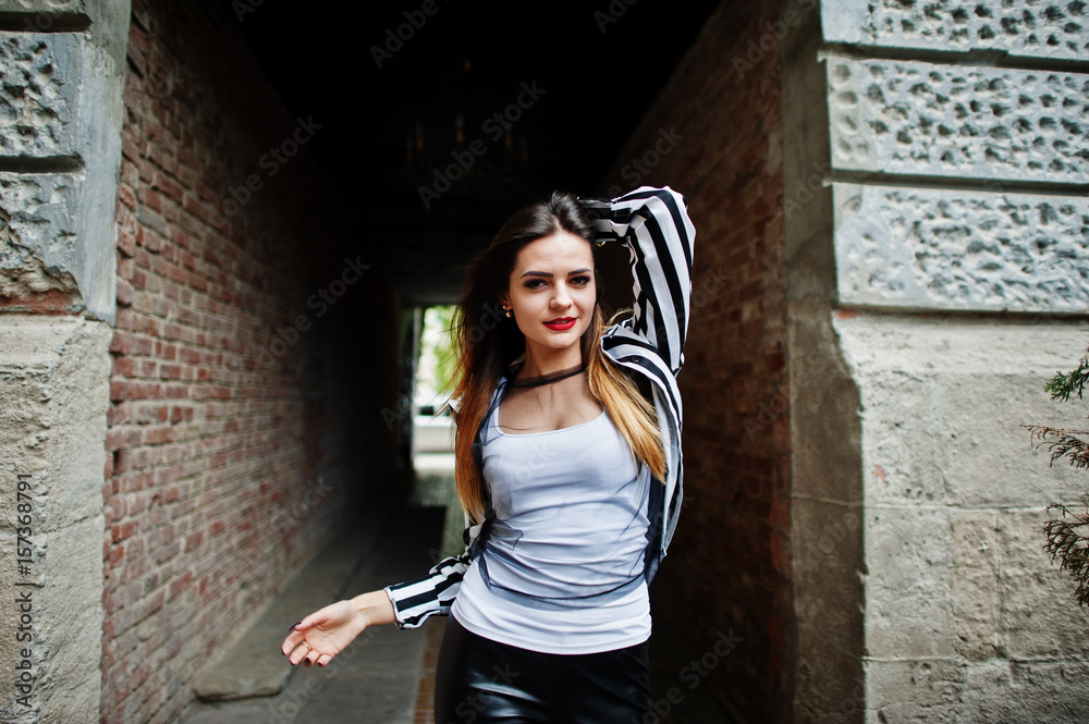 Fashionable woman look with black and white striped suit jacket, leather pants, posing at old street red brick tunnel. Concept of fashion girl.