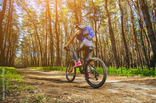 One young woman - an athlete in a helmet riding a mountain bike outside the city, on the road in a pine forest on a summer day.