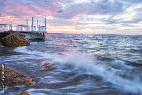 Dawn at the sea. White wooden pier on the shore and stones. The wave is approaching the shore. Long exposure.
