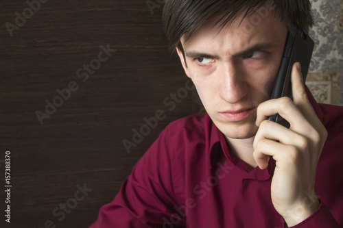 A young man in a purple shirt is waiting for an answer on a black smartphone, with an alarming suspicious look, frowning