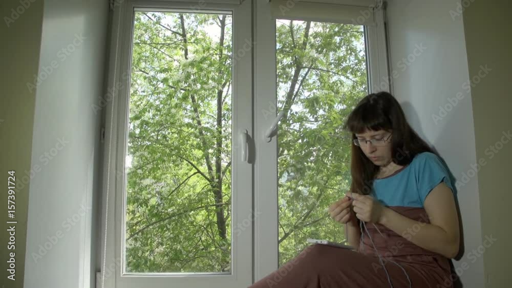 Young beautiful woman in glasses knits from blue yarn with knitting needles and uses a smartphone sitting on a windowsill at a bright spring day outside a tree with green leaves. Young beautiful woman in glasses knits from blue yarn with knitting needles and uses a smartphone sitting on a windowsill at a bright spring day outside a tree with green leaves.
