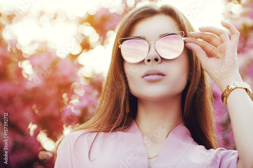 Outdoor close up portrait of young beautiful girl wearing stylish trendy glasses, posing in street, near blooming tree with pink flowers. Model looking up. Sunny day light. Female fashion concept
