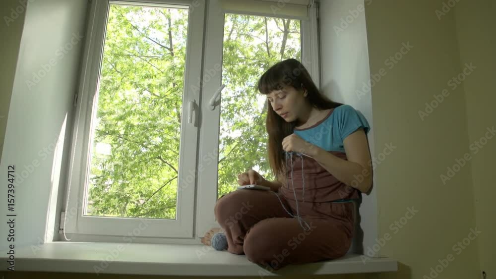 Young beautiful happy woman knits from blue yarn with knitting needles and uses smartphone sitting on window sill at bright spring day outside window trees with green leaves. Young beautiful happy woman knits from blue yarn with knitting needles and uses smartphone sitting on window sill at bright spring day outside window trees with green leaves.