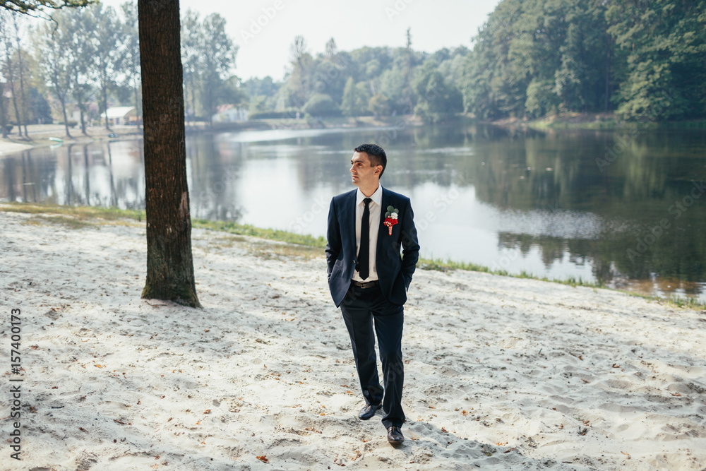 Groom in black suit with red boutonniere walks along the shore Groom in black suit with red boutonniere walks along the shore