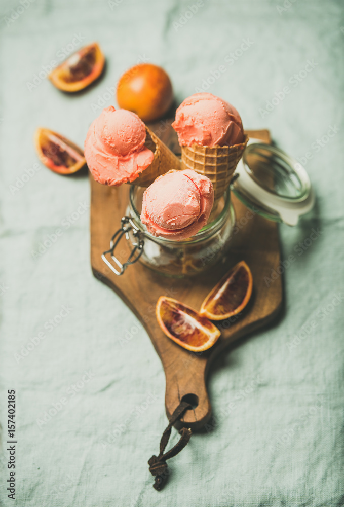 Refreshing summer blood orange ice cream or sorbet scoops in sweet waffle cones in glass jar served with fresh orange slices on rustic wooden board over light grey towel background, selective focus - obrazy, fototapety, plakaty 