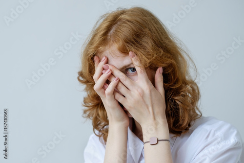 A red-haired girl with curls in embarrassment covers her face with her hands and looks through her fingers. Social phobia of the younger generation. photo