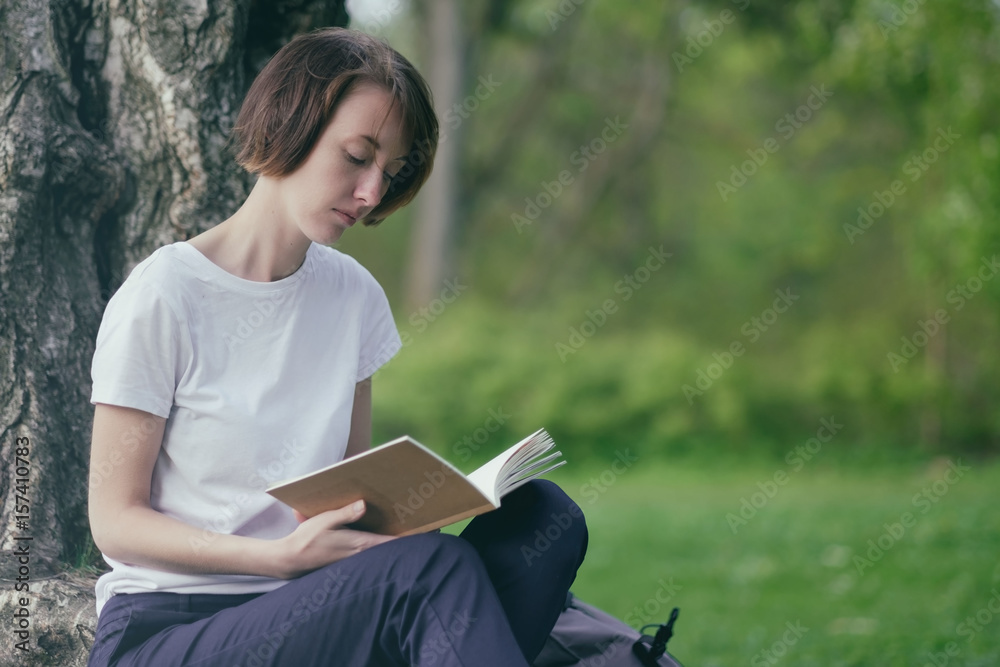 Obraz premium A girl reads a book in a park sitting by a tree on green grass. Relax on the outdoor summer