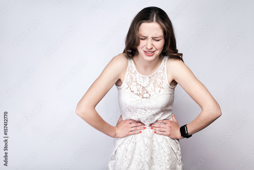 Portrait of beautiful woman with freckles and white dress and smart watch with stomach pain on silver gray background. healthcare and medicine concept.