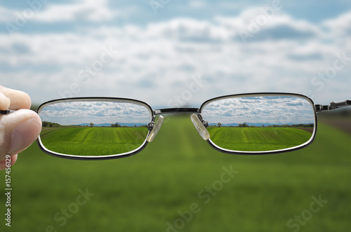 out of focus nature with farmlands and a hand holding a glasses that correct the vision