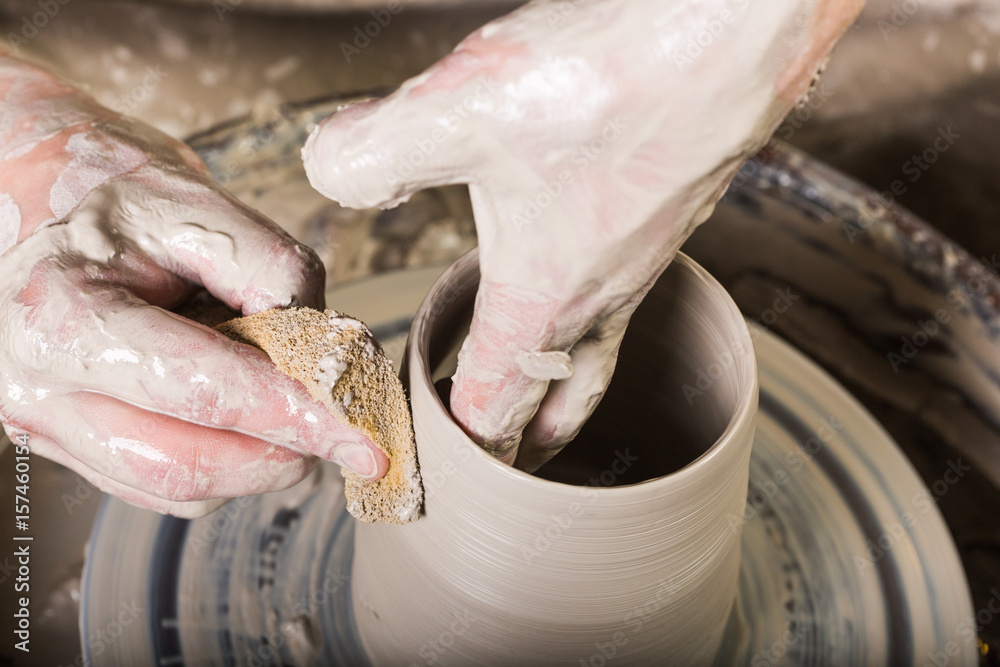 pottery, workshop, ceramics art concept - closeup on male fingers sculpt new utensil with a sponge and water, man's hands working with potter's wheel and raw fire clay, top view