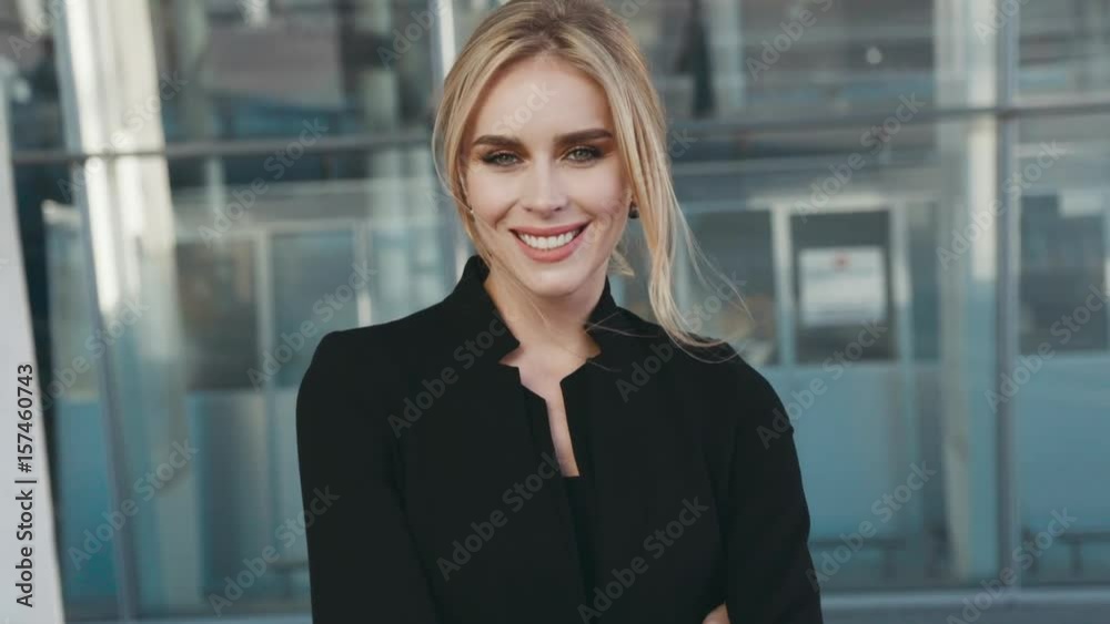 Beautiful blonde woman in a stylish black outfit, fashionable accessories, and natural makeup stands outside the airport terminal (business centre), looks right towards the camera and smiles brightly. Beautiful blonde woman in a stylish black outfit, fashionable accessories, and natural makeup stands outside the airport terminal (business centre), looks right towards the camera and smiles brightly.