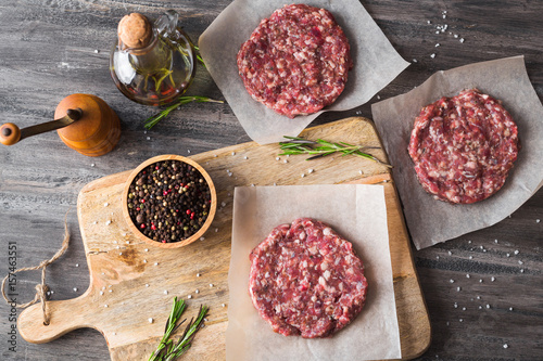 On a wooden cutting board on kraft paper there are raw beef burgers for burgers, a bottle of olive oil, spices, rosemary, a pepper mill, a cooking process, a top view