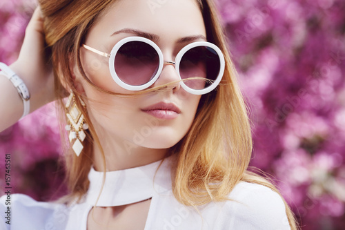 Outdoor close up portrait of young beautiful fashionable girl posing in street, near blooming tree with pink flowers. Model looking aside, wearing stylish glasses, wrist watch. Copy, empty space

