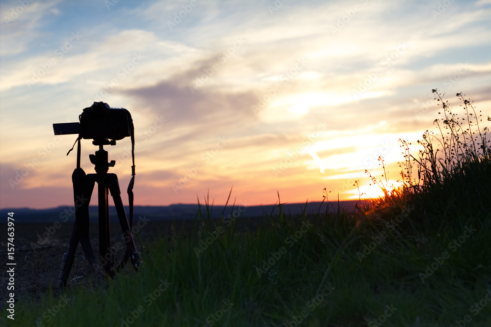 A camera set on a tripod aimed at a silhouette of a landscape at sunset. 