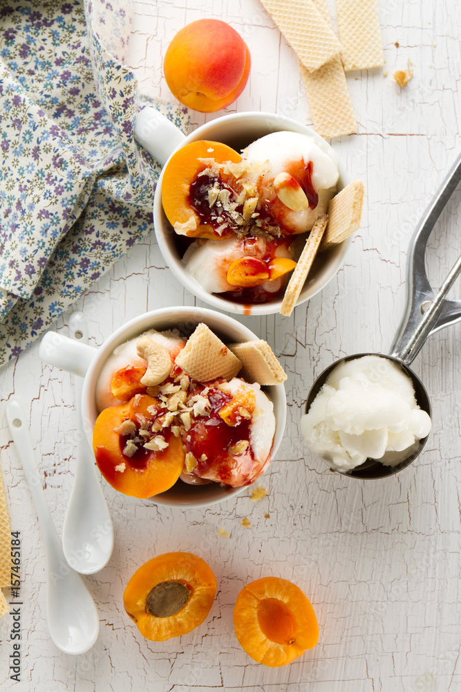 Tasty fresh cold appetizing vanilla ice cream with nuts, apricots, waffles and syrup on white table with ingredients for making dessert. Top View. Tasty fresh cold appetizing vanilla ice cream with nuts, apricots, waffles and syrup on white table with ingredients for making dessert. Top View.